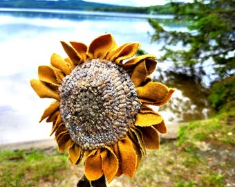 May include: A handcrafted sunflower sculpture featuring golden-yellow fabric petals arranged in multiple layers around a textured, multi-toned brown and grey center. The flower is attached to a thin stem with two dark brown felt leaves.