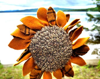 May include: A handcrafted sunflower decoration featuring a textured, dark brown, woven fabric center surrounded by layered petals made of solid mustard yellow felt and brown plaid wool fabric, attached to a thin, dark green felt-covered stem.