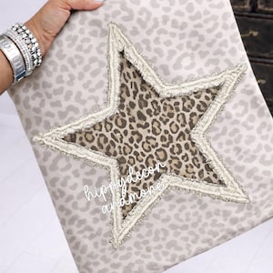 May include: A cushion featuring a light grey leopard print pattern. A large star with a leopard print centre and fringed edges is sewn onto the cushion. The text "hippy decor and more" is visible on the cushion.
