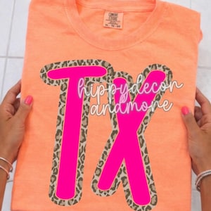 May include: An orange t-shirt with the text "Tx hippydecor and more" in a bright pink font with a leopard print border. The shirt is folded and held by hands with pink nail polish.