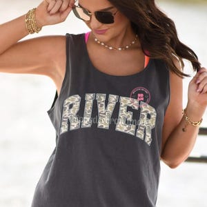 May include: Dark gray tank top with the word "RIVER" in a camouflage pattern across the front. The tank top has a round neckline and a sleeveless design. A small pink and white circular graphic is above the word "RIVER".