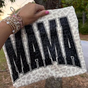 May include: A white t-shirt with a gray leopard print background and the word "MAMA" in large black block letters. The letters have a white outline. The shirt is being held up by a person.