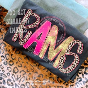 May include: Black t-shirt with the word "RAMS" in a combination of leopard print, pink, and gold lettering. The shirt is folded with other shirts in the background. Text on the shirt includes "Hippy Decor and More" and "Do Not Share Images."