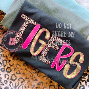 May include: Black t-shirt with the word "TIGERS" in a layered design. The letters are outlined in pink and gold, with a leopard print pattern on some letters. The text "DO NOT SHARE MY IMAGES" is also visible.