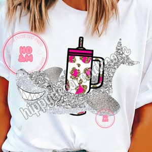 Puede incluir: Camiseta blanca con un diseño de tiburón brillante y una taza de café. La taza de café tiene un estampado de leopardo rosa y dorado. La camiseta tiene el texto "hippydeco".