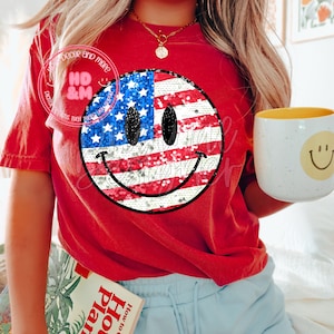 Puede incluir: Camiseta roja con un diseño de cara sonriente con lentejuelas que representa la bandera estadounidense. La camiseta la lleva puesta una persona que lleva pantalones cortos de mezclilla azul.