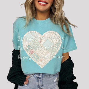 Könnte beinhalten: Ein hellblaues T-Shirt mit einem herzförmigen Design aus Patchwork-Stoff mit floralen und Spitzen-Details. Der Text "hippydecorandmore" wiederholt sich über das Design. Das Shirt wird mit einer Jeans und einer dunkelgrünen Strickjacke getragen.