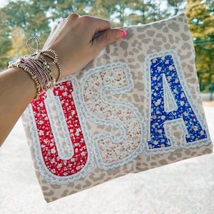 May include: A rectangular, light-coloured fabric with a leopard print pattern. The word "USA" is appliquéd on the fabric in red, floral, and blue patterned letters with a white lace trim.