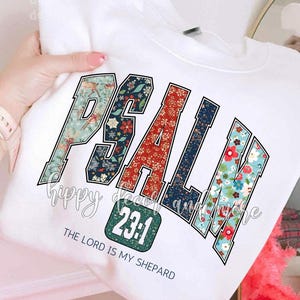 May include: White sweatshirt featuring the word "PSALM" in a floral design with black outlines. The number "23:1" is in a green square, with the text "THE LORD IS MY SHEPHERD" below. The sweatshirt is held by a person.