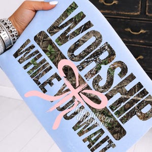 May include: A light blue sweatshirt with the words "WORSHIP WHILE YOU WAIT" in a camouflage pattern. A pink bow graphic is centered below the text. The sweatshirt has the text "hippydecor andmore" and other text at the bottom.