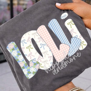 May include: Dark gray t-shirt with the word "lolli" in a patchwork design. The letters feature floral, gingham, and striped patterns, with bow accents. The text "hippy and more" is printed below the word "lolli".
