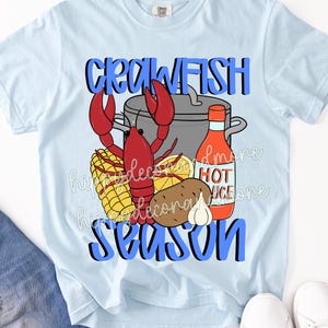 May include: Light blue t-shirt with a graphic featuring a red crawfish, a pot, corn, potato, garlic, and a bottle of hot sauce. The words "CRAWFISH SEASON" are in blue block letters.