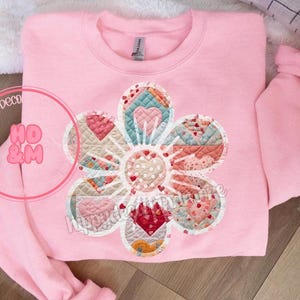 Puede incluir: Una sudadera rosa con un gran diseño de flor de patchwork colorido. La flor está hecha de cuadrados y triángulos de diferentes colores y tiene un contorno blanco.