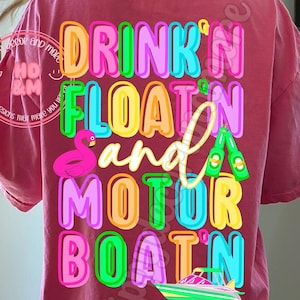 May include: A pink t-shirt with the text "DRINK'N FLOATIN and MOTOR BOATIN" in rainbow colors. The text is in a bubble font style. There is a pink flamingo float, a green and white motorboat, and a bottle of beer in the design.