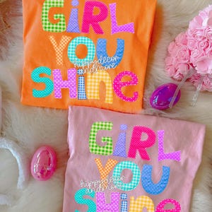 May include: Two colorful t-shirts with the words "GIRL YOU SHINE" in various patterned letters. One shirt is orange, the other is pink. Decorative Easter eggs and a pink flower bouquet are also in the image.