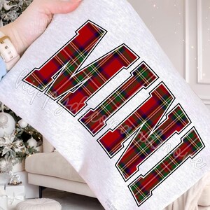 May include: A white pillow with the word "MINE" in large, bold letters. The letters are in a red, green, and yellow plaid pattern with a black outline. The pillow is being held in a person's hand.