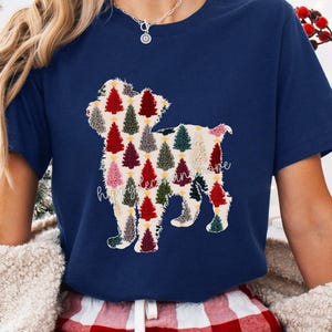 May include: Navy blue t-shirt featuring a dog-shaped design filled with colorful Christmas trees. The text "happy decorations and more" is written in cursive below the dog. The shirt is paired with red and white plaid pajama pants.