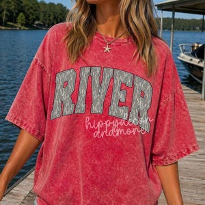 May include: Red, short-sleeved t-shirt with the word "RIVER" in a grey, textured font across the chest. Below the word "RIVER" is the phrase "hippy and more" in a cursive font. The shirt has a vintage, washed look.