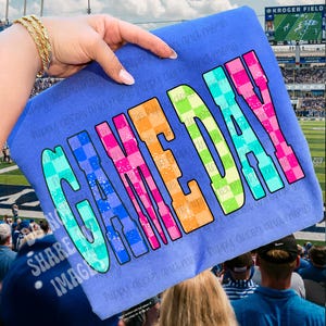 Puede incluir: Una camiseta azul con las palabras "GAME DAY" en letras grandes y coloridas a cuadros. La camiseta se sostiene frente a un estadio de fútbol, lo que sugiere un artículo de temática deportiva.