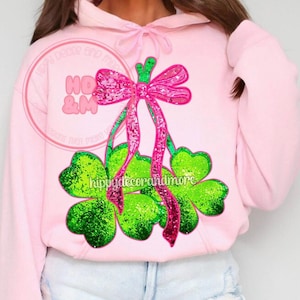 May include: A light pink hoodie with a sequined design of a pink bow and green shamrocks. The hoodie has a drawstring hood and long sleeves. The text "HO & M" and "hippydecorandmore" are visible on the design.
