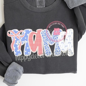Op de afbeelding: Een donkergrijs sweatshirt met het woord "MAM" in grote, kleurrijke letters. De letters zijn gemaakt van stofresten in rood, wit en blauw met een patchwork-ontwerp. De letters zijn wit omrand.