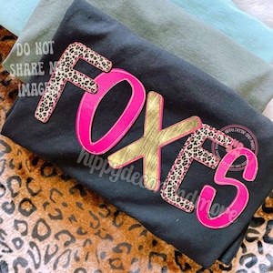 May include: Black t-shirt with the word "FOXES" in large, layered letters. The letters have a pink outline, gold and leopard print accents. The shirt is folded, with other shirts visible in the background.