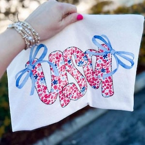 Op de afbeelding: Wit T-shirt met de letters "USM" in een bloemenpatroon van rode en blauwe bloemen. De letters zijn zwart omlijnd en versierd met blauwe strikken. Het T-shirt wordt door een hand vastgehouden.
