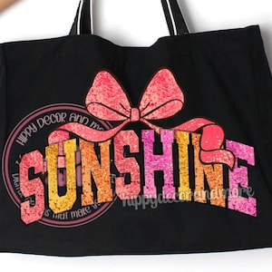 May include: Black canvas tote bag featuring a pink and orange glitter design that reads "Sunshine" with a pink bow on top. The text "Hippy Decor and More" is written around the design.