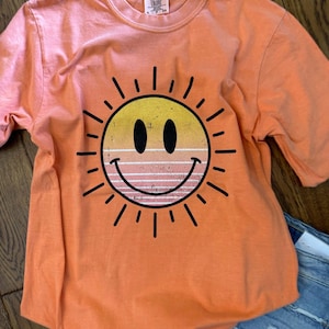 May include: Orange t-shirt featuring a large, smiling sun graphic. The sun has a yellow and orange gradient with black outlines and rays. The shirt is made of a soft material and has short sleeves.