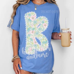 May include: A light blue t-shirt with a floral letter "R" design and the text "happy decor and more". The floral design includes pink, blue, and green flowers. The shirt is a comfortable, casual style.