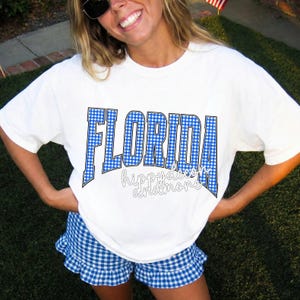 Puede incluir: Camiseta blanca con la palabra "FLORIDA" en estampado de cuadros vichy azules. La camiseta también presenta el texto "hippydeco andmore". La persona de la imagen lleva unos pantalones cortos a juego con cuadros vichy azules.
