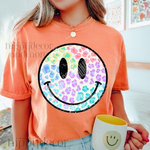 Puede incluir: Una camiseta de color melocotón con un gráfico de cara sonriente de arcoíris y estampado de leopardo. La camiseta es de corte holgado y tiene cuello redondo.