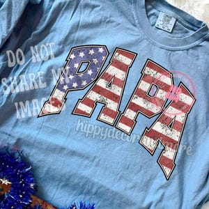 Puede incluir: Una camiseta de manga larga azul claro con la palabra "PAPA" impresa en rojo, blanco y azul, que se asemeja a la bandera estadounidense. La camiseta tiene un aspecto desgastado, lo que le da un toque vintage.
