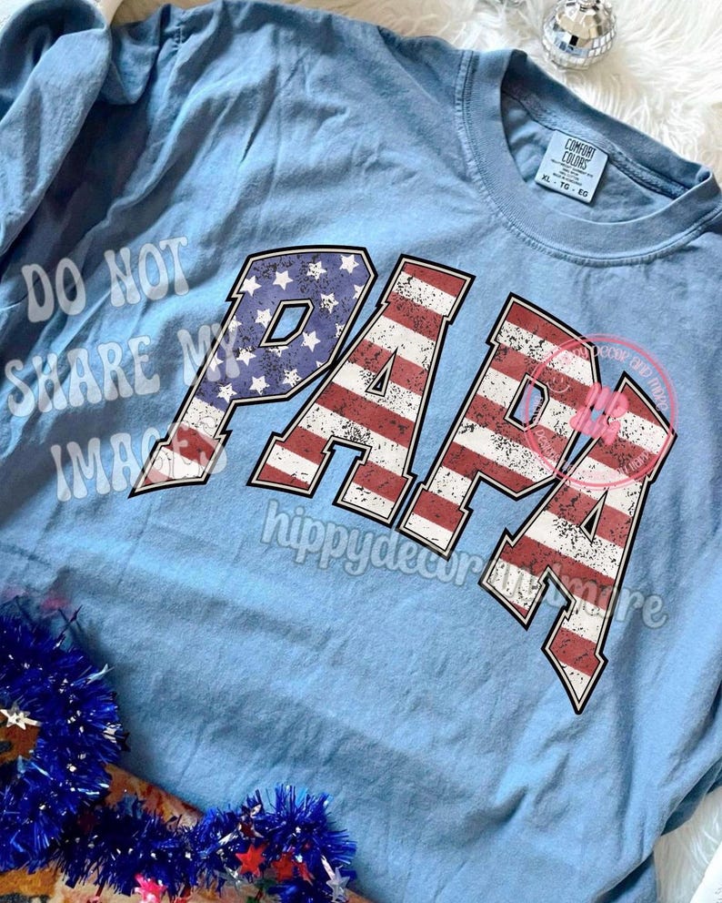 Puede incluir: Una camiseta de manga larga azul claro con la palabra "PAPA" impresa en rojo, blanco y azul, que se asemeja a la bandera estadounidense. La camiseta tiene un aspecto desgastado, lo que le da un toque vintage.