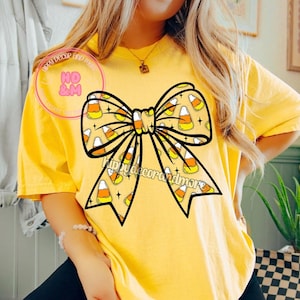 May include: Yellow t-shirt featuring a large bow design filled with candy corn and small stars. The bow has a black outline. The text "Hippy decor and more" is printed on the shirt. A necklace and bracelet are also visible.