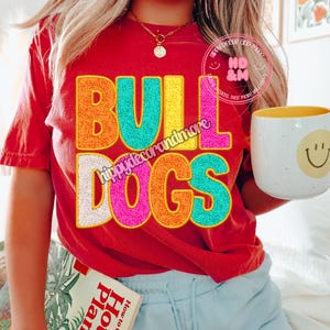 Op de afbeelding: Een rood T-shirt met de woorden "BULLDOGS" in kleurrijke, pluche letters. De letters zijn zwart omrand. Het shirt wordt gedragen door een persoon met blond haar en een gouden ketting.
