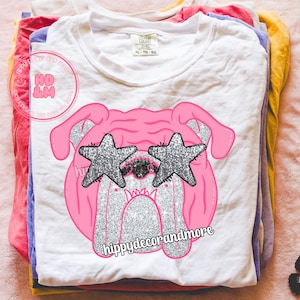 May include: White XL t-shirt featuring a pink bulldog design wearing glitter star-shaped sunglasses. The bulldog has silver glitter accents. The text "hippydecorandmore" is printed below the design. The shirt is stacked with other colorful shirts.