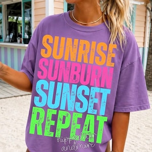 May include: A purple t-shirt with the words "SUNRISE SUNBURN SUNSET REPEAT" in orange, pink, blue, and green block letters. The shirt also has the text "hippydecor andmore" in a smaller font. The shirt is short-sleeved.