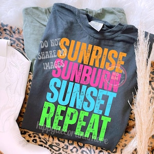 May include: A dark gray t-shirt with the words "Sunrise, Sunburn, Sunset, Repeat" in orange, pink, blue, and green. The shirt also has the text "Do Not Share My Images". A pair of white cowboy boots and a green shirt are also visible.