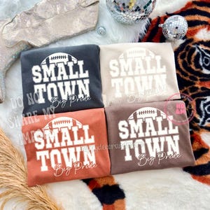 May include: Four sweatshirts in various colors, each featuring the text "SMALL TOWN Big Pride" with a football graphic. The sweatshirts are in shades of gray, tan, rust, and brown. The image includes a disco ball and a pair of sparkly boots.