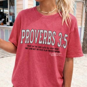 Puede incluir: Una camiseta roja con el texto "PROVERBIOS 3.5" en blanco, con puntos. Debajo, se lee "TRUST IN THE LORD WITH ALL YOUR HEART AND LEAN NOT ON YOUR OWN UNDERSTANDING". La camiseta también tiene el texto "hippydecor andmore".