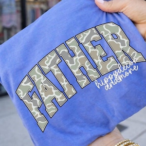 Peut inclure: Un t-shirt bleu avec le mot "FATHER" en grandes lettres en blocs avec un motif camouflage. Le texte "hippydecor andmore" est &eacute;crit en dessous du mot "FATHER" dans une police cursive. Le t-shirt est tenu par une personne.