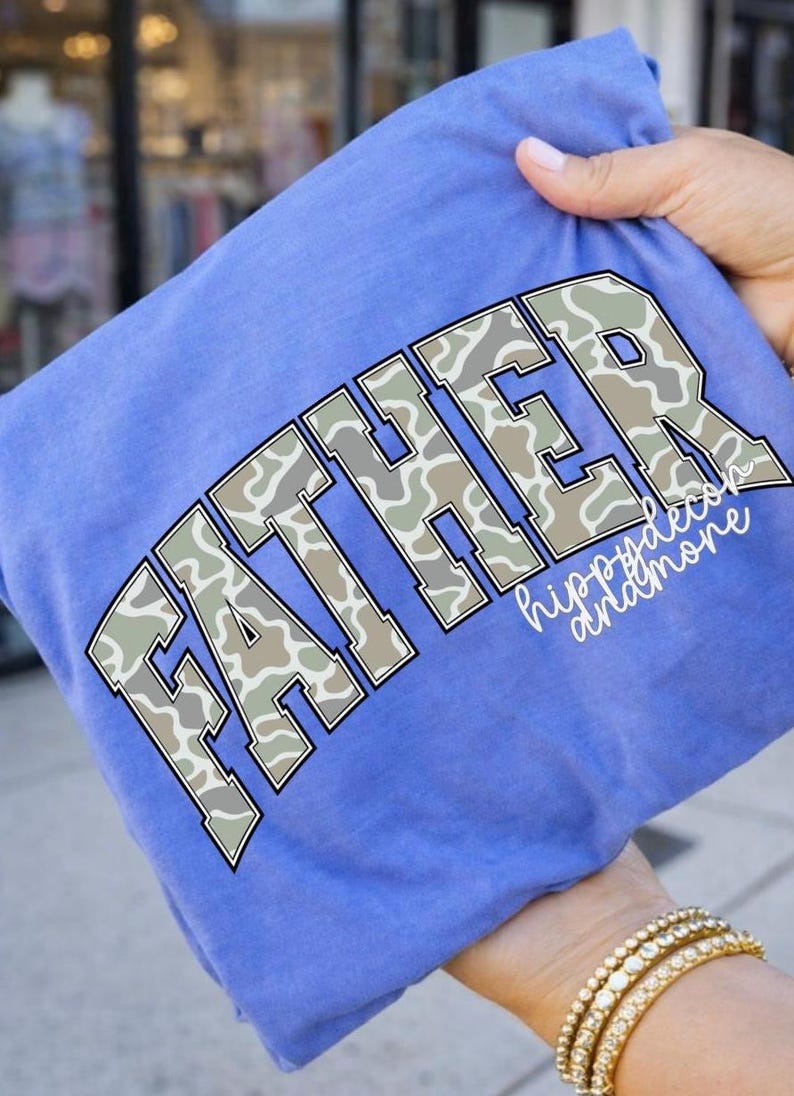 Peut inclure: Un t-shirt bleu avec le mot "FATHER" en grandes lettres en blocs avec un motif camouflage. Le texte "hippydecor andmore" est &eacute;crit en dessous du mot "FATHER" dans une police cursive. Le t-shirt est tenu par une personne.