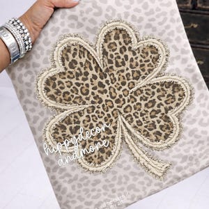May include: A decorative pillow with a large, leopard print shamrock design. The shamrock is outlined with a fringed trim and set against a light gray background with a subtle leopard print pattern. The text "hippydecon and more" is visible.