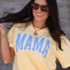 May include: A light yellow t-shirt with the word "MAMA" in large, light blue letters with white polka dots. The shirt is short-sleeved and has a relaxed fit. The text "happydecorn andmore" is printed on the shirt.
