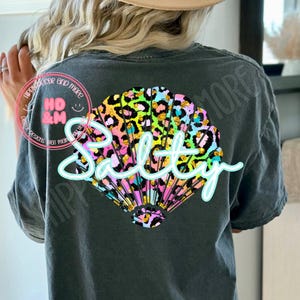 May include: Dark grey t-shirt with a colourful seashell design. The seashell features a leopard print pattern in shades of pink, yellow, green, and blue. The word "Salty" is written across the shell in a light blue script font.