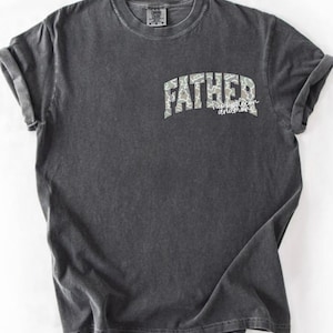 Peut inclure: T-shirt gris fonc&eacute; avec des manches retrouss&eacute;es. Le t-shirt affiche le mot "FATHER" dans un motif camouflage, arqu&eacute; sur la poitrine, avec les mots "happiest son and more" en dessous dans une police script. Le t-shirt a un aspect vintage et us&eacute;.