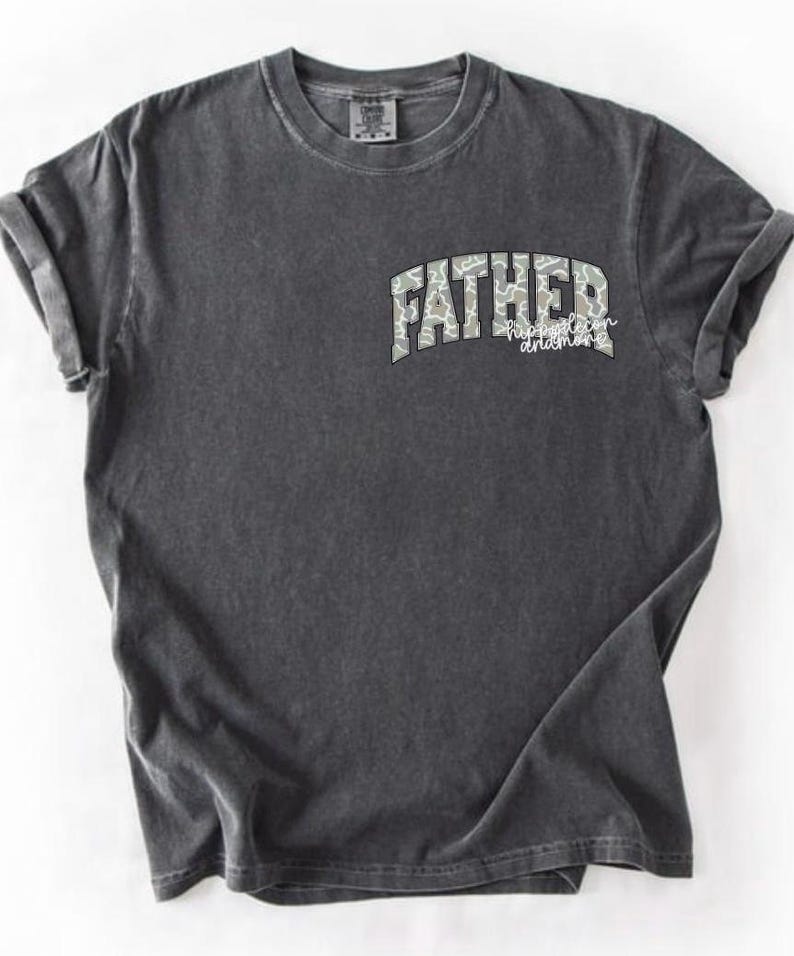 Peut inclure: T-shirt gris fonc&eacute; avec des manches retrouss&eacute;es. Le t-shirt affiche le mot "FATHER" dans un motif camouflage, arqu&eacute; sur la poitrine, avec les mots "happiest son and more" en dessous dans une police script. Le t-shirt a un aspect vintage et us&eacute;.