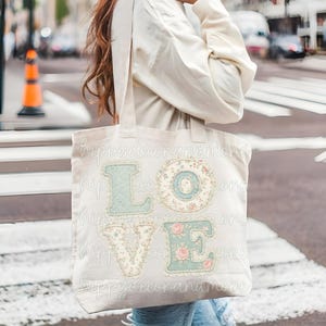 Pode incluir: Uma sacola tote bege com a palavra "LOVE" aplicada na frente. As letras são em estilo patchwork com detalhes florais e de renda em tons de azul, rosa e creme. As palavras "happydecorandmore" estão impressas na sacola.