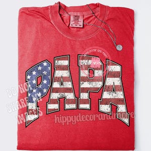 Puede incluir: Camiseta roja con un dise&ntilde;o de bandera estadounidense desgastado que deletrea "PAPA".